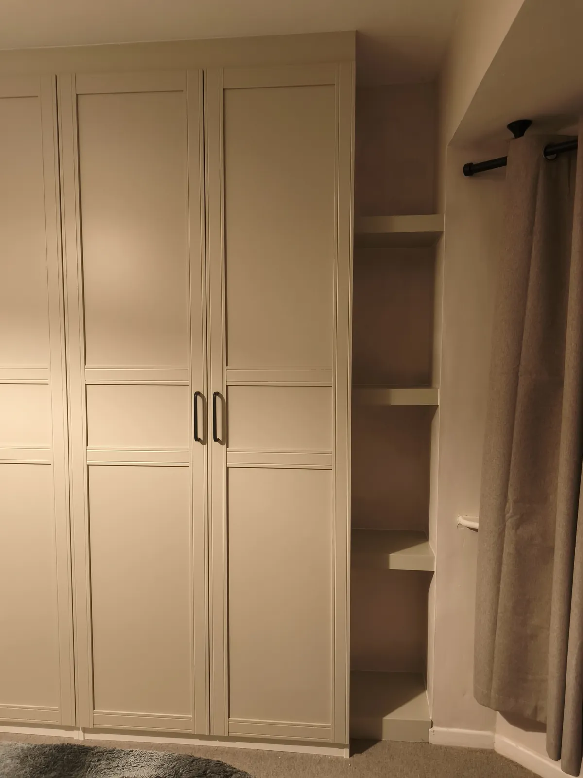 IKEA PAX wardrobes with beige shaker doors and black handles, fitted tight to the wall and ceiling, with open shelves in the recess beside them